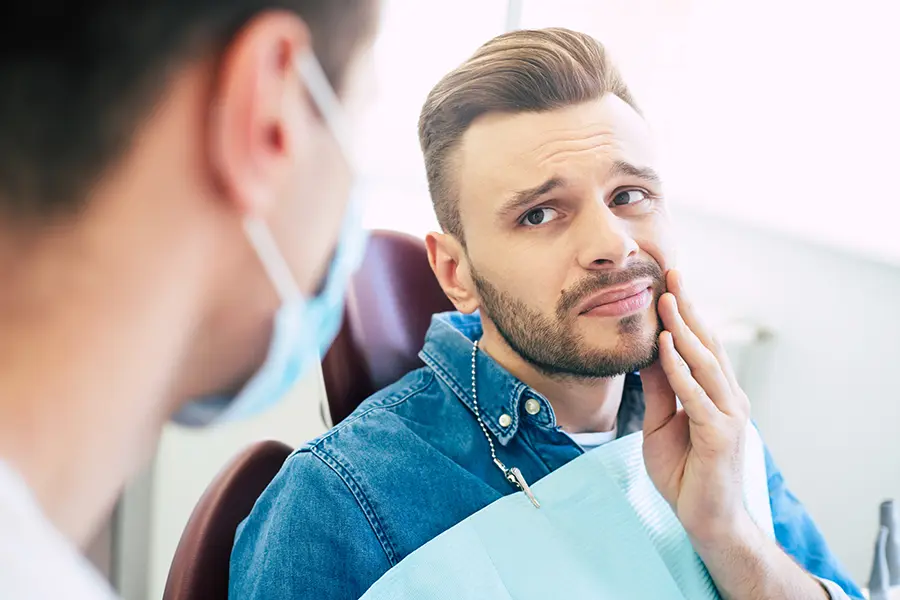 A man with a worried face is holding his hand on his cheek because of irritating pain in front of a dentist